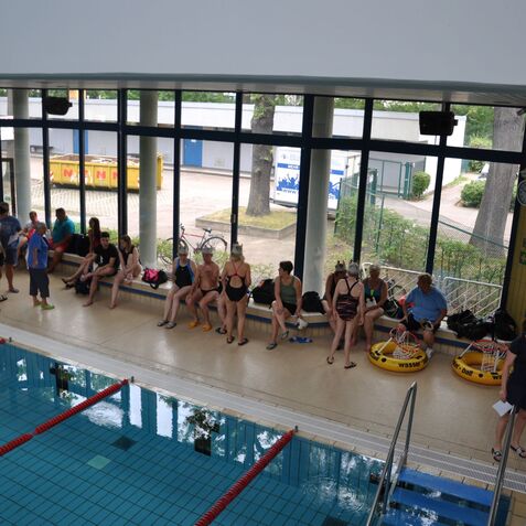Sitzende Schwimmer in Badebekleidung an einer Schwimmhalle, mit Wasser im Vordergrund und gro&szlig;en Fenstern im Hintergrund.