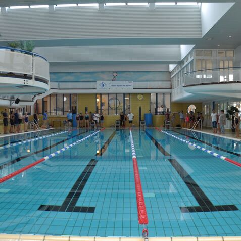 Schwimmbecken mit klar wasser, Schwimmern an der Seite und einer Zuschauerterrasse im Hintergrund.