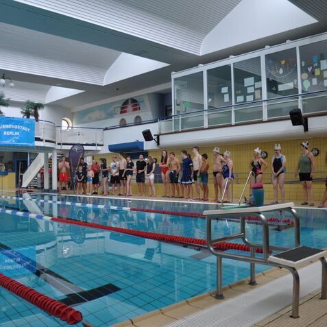 Schwimmerinnen und Schwimmer stehen am Beckenrand eines hallenbad, bereit f&uuml;r einen Wettkampf unter dem Banner &bdquo;SC Siemensstadt Berlin&ldquo;.