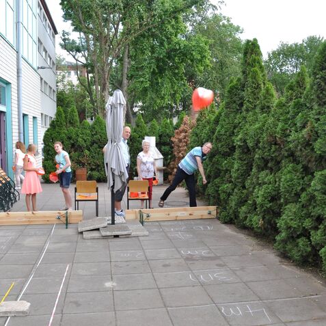 Gruppenaktivit&auml;t im Freien: Kinder und Erwachsene spielen mit einem roten Ball zwischen Hecken und Holzplattformen.