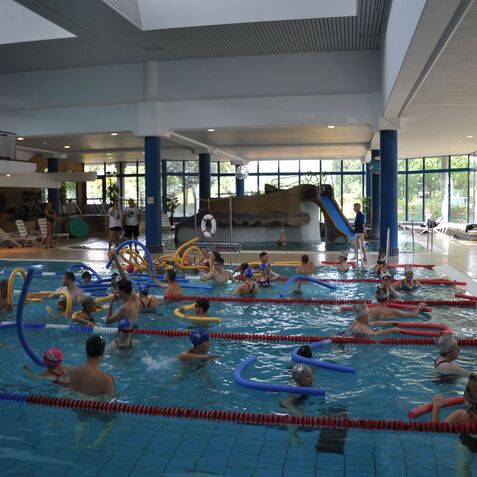 Gruppenschwimmen mit Kindern im Pool, umgeben von Nudelspielzeugen und Rutschen, in einer hellen Halle mit Fenstern.