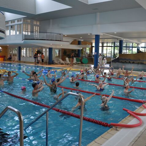 Gruppenschwimmen von Kindern in einem Hallenbad, die &Uuml;bungen im Wasser durchf&uuml;hren, mit Wellenbrechern und Rutschen im Hintergrund.