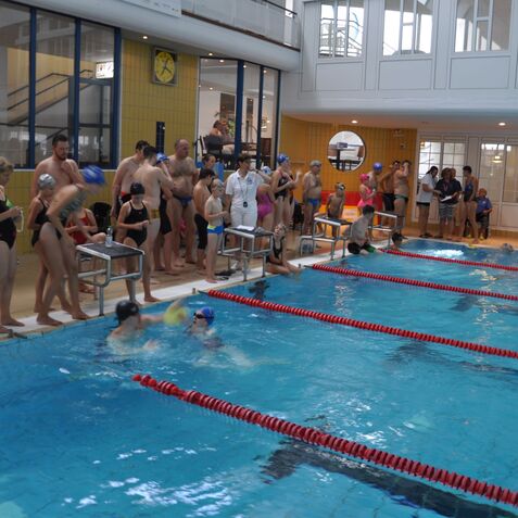 Schwimmwettkampf mit Teilnehmern am Beckenrand und Wettk&auml;mpfern im Wasser in einem Hallenbad.