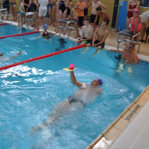 Ein Schwimmer liegt auf dem R&uuml;cken im Wasser und h&auml;lt einen pinken Becher auf einem Teller &uuml;ber dem Kopf.