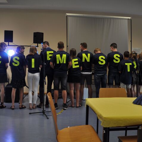 Gruppe von Personen mit T-Shirts, auf denen gro&szlig;e Buchstaben stehen, in einer Veranstaltungshalle mit St&uuml;hlen und Tisch.