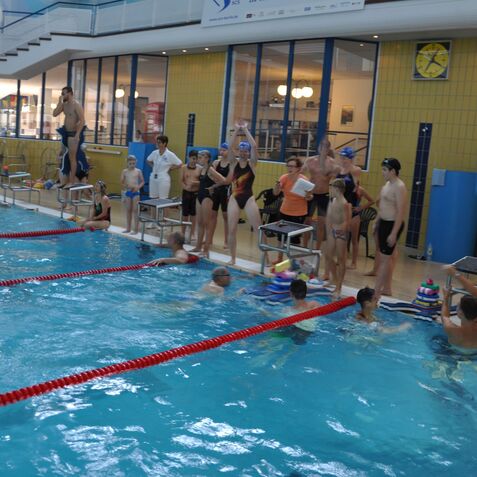 Schwimmwettkampf mit Teilnehmern am Beckenrand und im Wasser in einem Hallenbad, einige jubeln, andere bereiten sich vor.