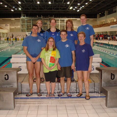 Gruppe von Schwimmern in blauen Shirts steht am Beckenrand eines Schwimmbads mit Wettkampfbecken im Hintergrund.