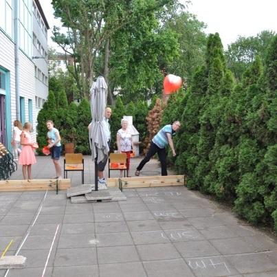 Szenenbild einer Gartenveranstaltung mit Personen, die an einer Spielstation mit Holzlatten und einem roten Ball teilnehmen.