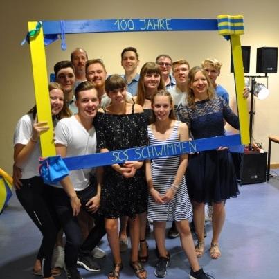 Gruppenfoto von zwölf Personen in festlicher Kleidung mit einem Rahmen, der "100 Jahre SC Schwimmen" zeigt.