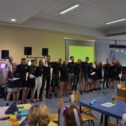 Gruppe von Personen in schwarzer Kleidung stehen zusammen auf einer Bühne, Umrahmung mit Tischen und Gästen im Hintergrund.