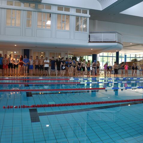 Gruppierung von Schwimmern am Beckenrand bei einem Wettkampf in einem Hallenbad, auf der linken Seite eine Trainerin.
