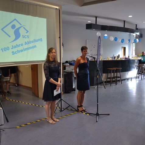Zwei Frauen sprechen auf einer Veranstaltung vor einer Leinwand mit dem Motto „100 Jahre Abteilung Schwimmen“.