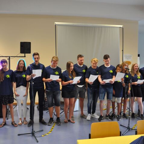 Gruppe von Jugendlichen singt auf einer Bühne, während sie Texte in der Hand halten. Mikrofone und Lautsprecher im Hintergrund.