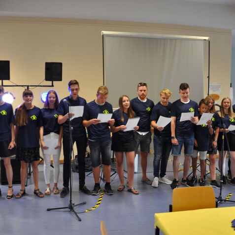 Gruppe von Jugendlichen in blauen T-Shirts singt auf einer Bühne, einige halten Blätter in der Hand.