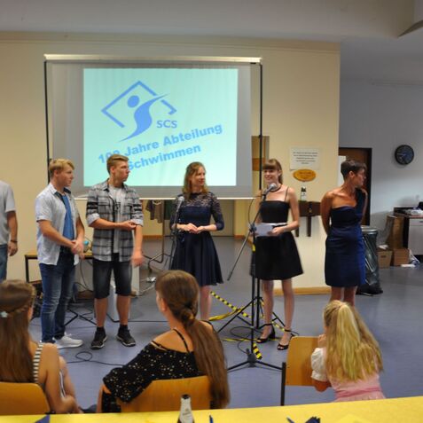 Gruppenszene bei einer Feier mit mehreren Personen am Mikrofon und einer Präsentation im Hintergrund.