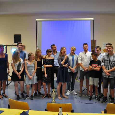 Gruppe von Menschen auf einer Veranstaltung, eine Person spricht, Pokal und Mikrofon sichtbar, bunte Lichter im Hintergrund.