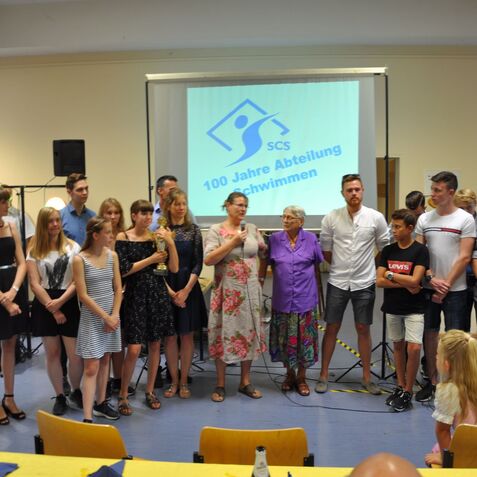 Gruppe von Personen bei einer Veranstaltung mit einer Präsentation zum 100-jährigen Jubiläum der Schwimmabteilung.