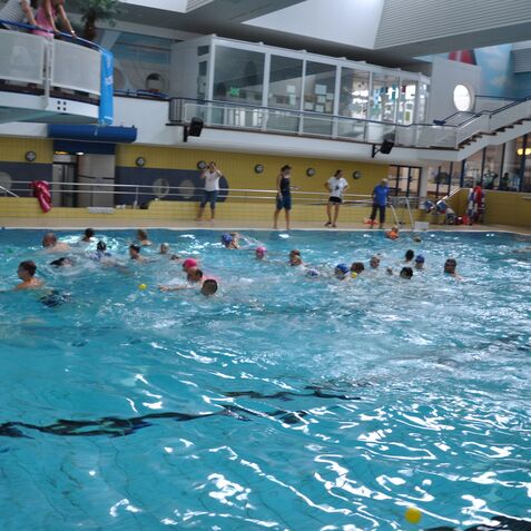 Gruppensport im Hallenbad mit mehreren Schwimmern, Spielzeugbällen und Aufsichtspersonal am Beckenrand.