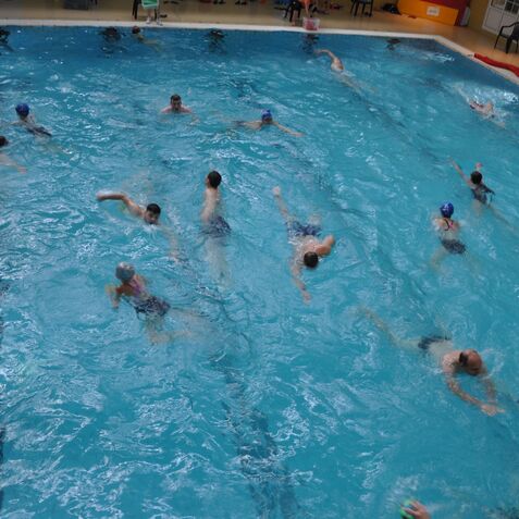 Gruppenschwimmen von Menschen unterschiedlichen Alters in einem Hallenbad mit blauer Wasseroberfläche.