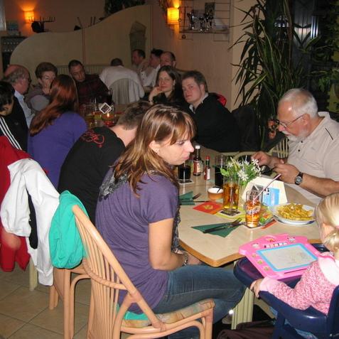 Gruppe von Personen an einem Tisch in einem Restaurant, einige essen, andere unterhalten sich, ein Kind malt.