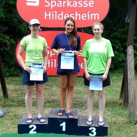 Drei Frauen stehen auf dem Siegerpodest mit Urkunden, umgeben von grüner Natur und Sponsorenfahnen.
