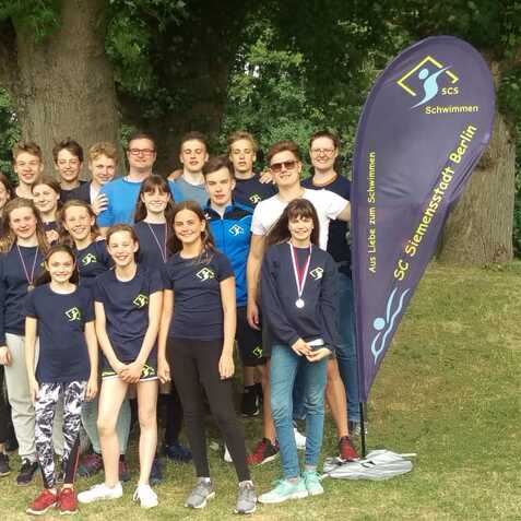 Gruppe von Schwimmern in blauen Shirts mit Vereinsbanner im Hintergrund, versammelt unter einem Baum im Freien.