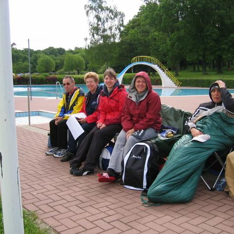 Fünf Personen sitzen angeordnet auf einer Bank am Schwimmbeckenrand, umgeben von Grünanlagen und Bäumen.