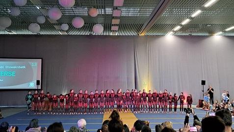Gruppenvorstellung von Sportlerinnen in schwarzen und roten Bodys auf einer Bühne mit applaudierendem Publikum im Hintergrund.