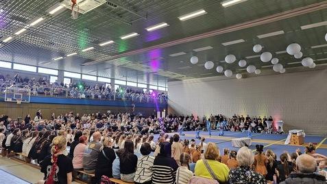 Große Menschenmenge in einer Sporthalle beobachtet eine Aufführung auf der Matte, umgeben von bunten Lichtern und Dekoration.