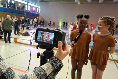 Ein Mädchen interviewt zwei Kinder in braunen Kostümen auf einer Turnbühne, während Zuschauer im Hintergrund stehen.