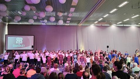 Bühne mit vielen Tänzern in bunten Outfits, die sich vor Publikum präsentieren; Bildschirm zeigt „Danke!“.