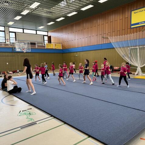 Gruppe von Kindern in rosa T-Shirts &uuml;bt auf Matten in einer Sporthalle, w&auml;hrend Trainerin anleitet.