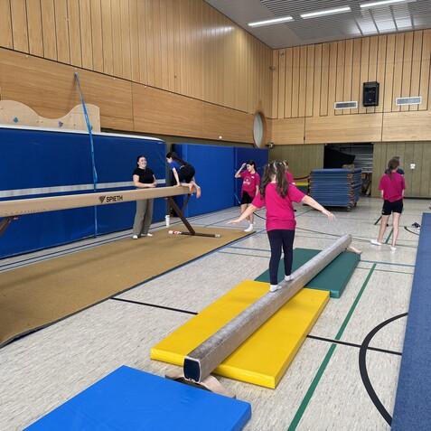 Sechs Kinder in pinken T-Shirts &uuml;ben Balancieren auf einem Schwebebalken in einer Turnhalle. Eine Trainerin beobachtet.