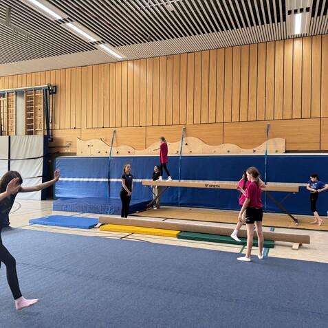Sechs Kinder &uuml;ben auf verschiedenen Ger&auml;ten in einer Turnhalle, darunter ein Schwebebalken und Matten.