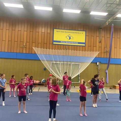 Gruppe von Kindern in T-Shirts mit den Worten „Sport Club“ beim Training in einer Sporthalle mit gymnastischen Geräten.