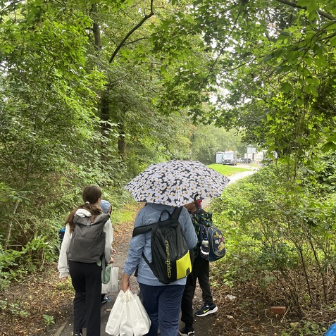 Gruppe von Personen mit Rucks&auml;cken, die unter einem Regenschirm einen schmalen Weg im Wald entlanggehen.