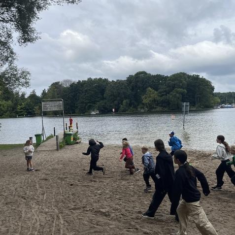 Gruppe von Kindern l&auml;uft am Sandstrand eines Sees, im Hintergrund ein Steg mit einer Mauer und B&auml;umen.