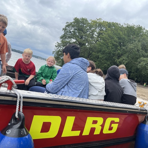 Auf einem Boot sitzen mehrere Kinder in lockerer Kleidung, mit einem See und B&auml;umen im Hintergrund.