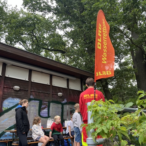 DLRG-Wasserrettung: Gruppe von Kindern und Betreuern vor einem Holzhaus, mit Flagge im Hintergrund und B&auml;umen.