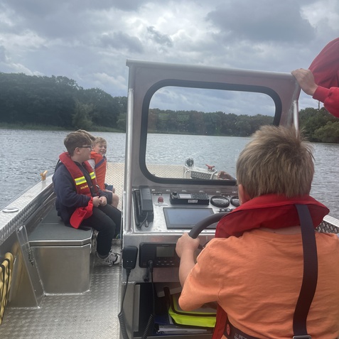 Vier Kinder in Rettungswesten fahren in einem Boot auf einemSee, w&auml;hrend ein Kind am Steuer sitzt.