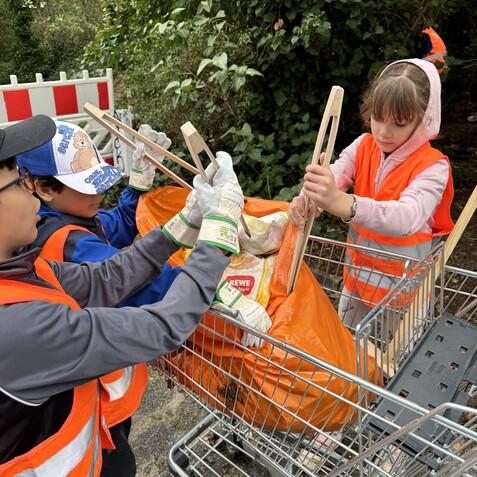 Kinder mit Handschuhen und Warnwesten sortieren Müll in einen Einkaufswagen, umgeben von Bäumen und Grün.