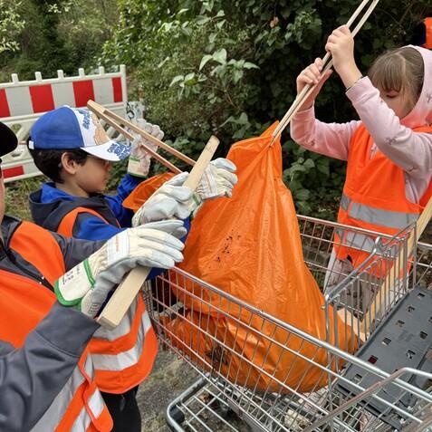 Kinder in orangenen Westen sammeln Müll in einem Einkaufswagen und verwenden Holzstangen, um Abfall in einen Sack zu legen.