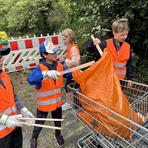 Vier Kinder in Warnwesten sammeln Müll mit Zangen und einem großen, orangefarbenen Sack neben einem Einkaufswagen.