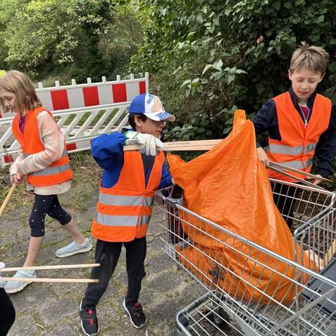 Kinder in Warnwesten sammeln Müll mit Greifern und einer leeren Einkaufskarre, umgeben von Bäumen und einer Baustellenabsperrung.