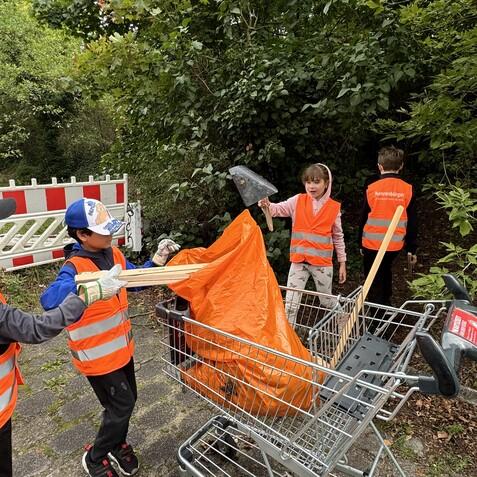 Kinder in Warnwesten sammeln Müll in einem Einkaufswagen, während sie mit orangefarbenen Müllsäcken und Werkzeug arbeiten.