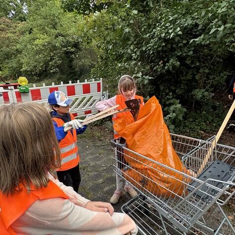 Vier Kinder in leuchtenden Westen sammeln Müll in einem Einkaufswagen im Freien, umgeben von Bäumen und Büschen.