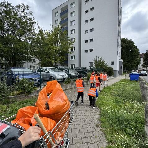Gruppe von Kindern in orangen Westen, die mit einem Einkaufswagen voll Müll einen Gehweg entlanggehen.
