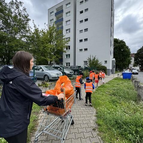 Gruppe von Kindern in orangefarbenen Westen sammelt Müll mit einem Einkaufswagen vor einem Wohngebäude.