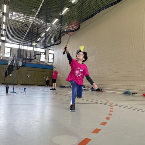Kind in pinkem T-Shirt schlägt mit einem Schläger einen Federball in einer Sporthalle. Im Hintergrund sind weitere Kinder sichtbar.