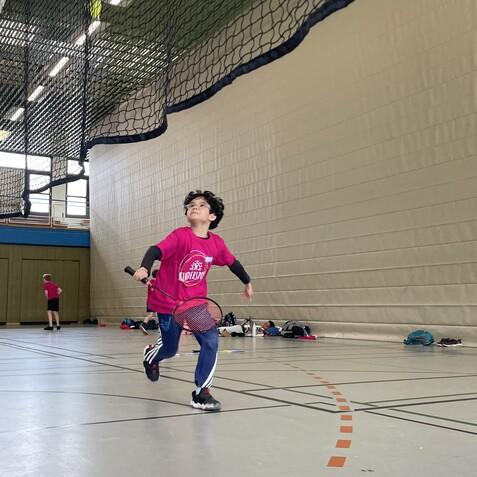 Junge in pinkem T-Shirt spielt Tennis, läuft mit Schläger in Richtung des hohen, fliegenden Balls in einer Halle.